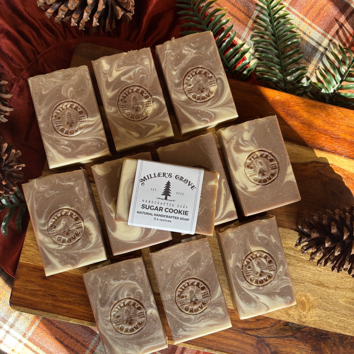 Nine bars of Miller's Grove handmade soap, featuring a central 'Sugar Cookie' bar with its paper label, arranged on a rustic wooden board with pinecones and holiday greenery.
