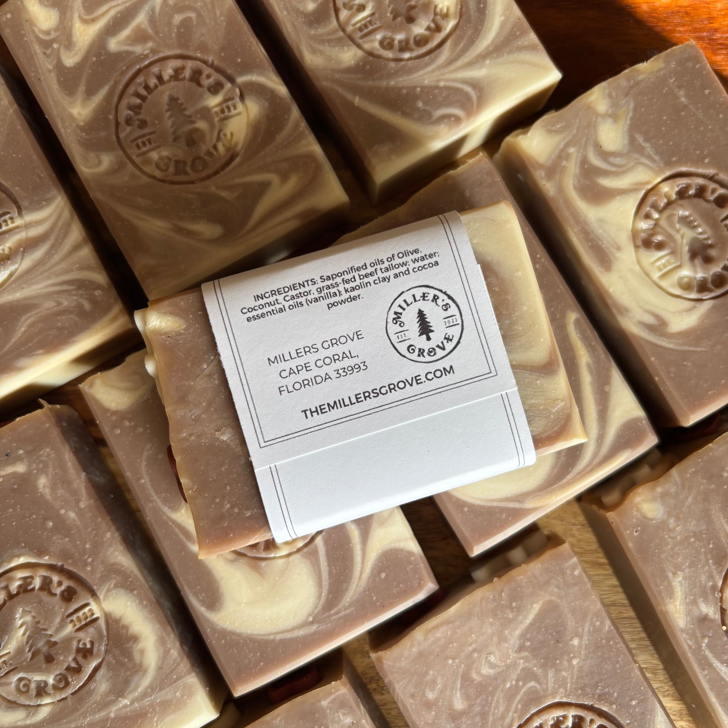 Several rectangular bars of soap with a marbled brown and cream pattern are arranged on a wooden surface. One soap bar has a white paper label wrapped around it with printed text and the Miller's Grove logo.