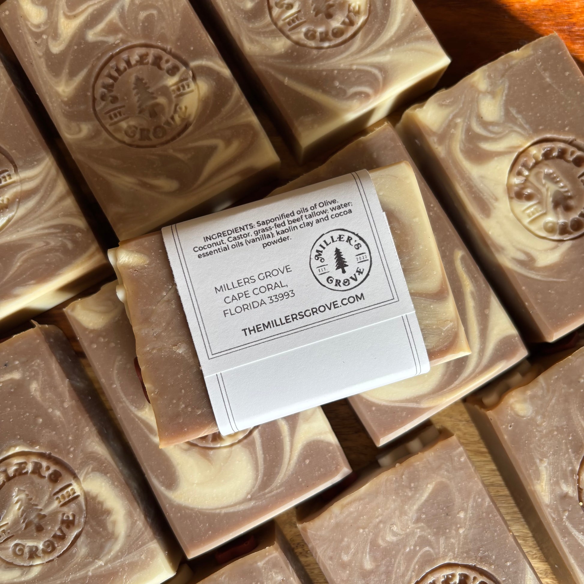 Several rectangular bars of soap with a marbled brown and cream pattern are arranged on a wooden surface. One soap bar has a white paper label wrapped around it with printed text and the Miller's Grove logo.
