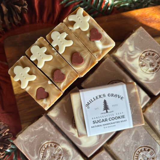 Handcrafted sugar cookie soap bars arranged on a wooden board, with one bar wrapped in a paper label reading Miller's Grove Handcrafted Soap Sugar Cookie. The soaps are decorated with white gingerbread men and red hearts, with other marbled brown and white soaps in the background.