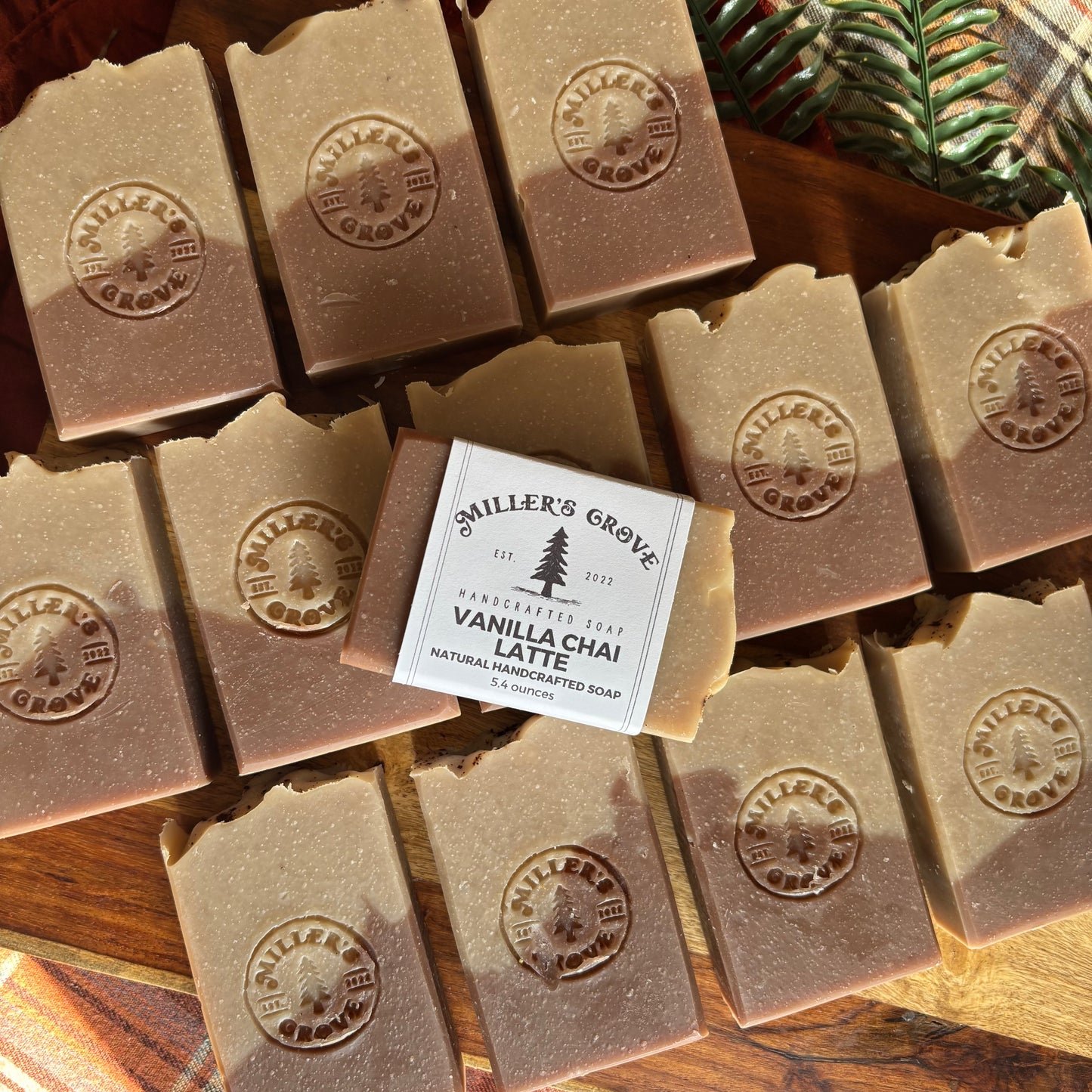 Several bars of Miller's Grove Vanilla Chai Latte handmade soap arranged on a wooden surface, with one central bar wrapped in a white paper label. The soaps are a warm light brown/tan color with an embossed circular logo, and a green and brown plaid fabric is partially visible in the top right corner.