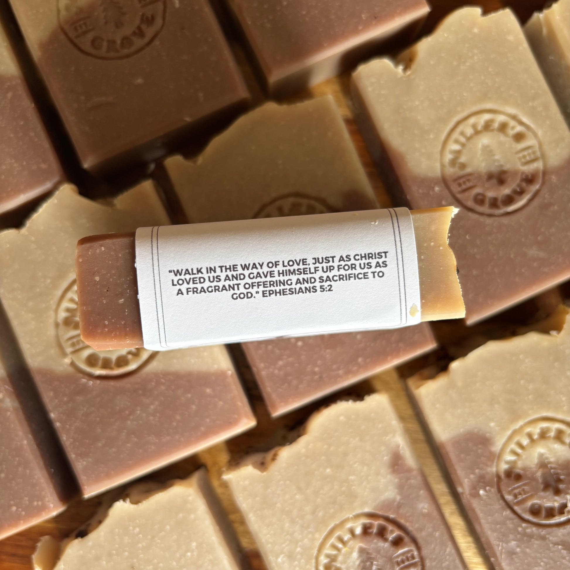 A close-up of a rectangular stick of butter soap lying horizontally on top of several stacked butter soaps. The central soap has a white paper label wrapped around it with printed black text, and the surrounding soaps are light tan and medium brown with embossed circular logos.