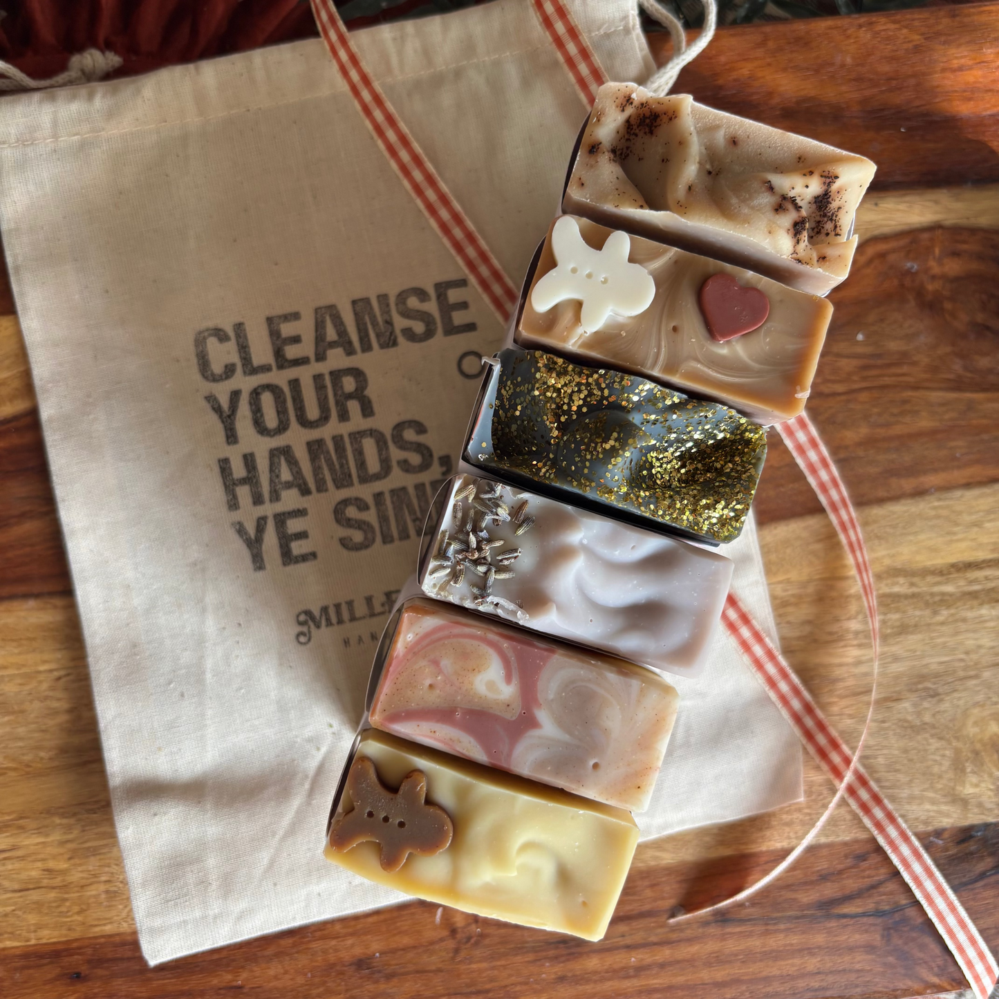Six artisanal handmade soaps in various designs and colors are stacked vertically on a beige cotton drawstring bag with black text, placed on a wooden table next to a red and white striped ribbon.