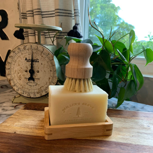 Beige rectangular brick of soap, on a wood soap tray with a scrubber brush on top of the soap